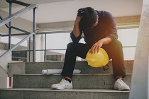 hombre sentado en escaleras con cabeza baja, sujetando casco amarillo